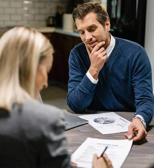 Hombre concentrado y pensativo en una mesa de cocina, revisando gráficos y documentos que le presenta una asesora (vista desde atrás), ilustrando el proceso de consulta y los servicios de planificación financiera o contable.