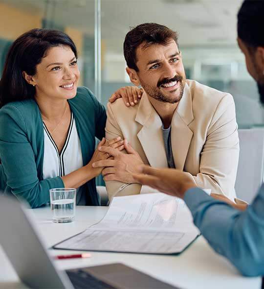 Pareja joven y sonriente sentada en una oficina, mirando atentamente y con tranquilidad a un asesor que les explica documentos, ilustrando la confianza que se obtiene al recibir una buena asesoría fiscal y financiera.